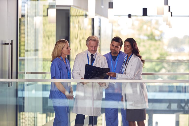 Multi-Cultural Medical Team Wearing White Coats And Scrubs Discuss Patient Scan Meeting In Hospital